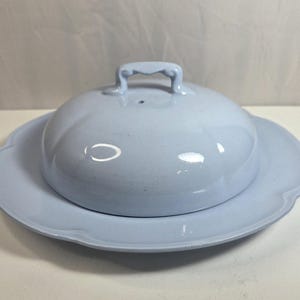 May include: A light blue ceramic butter dish with a matching domed lid. The base features a scalloped edge, while the lid has a smooth, rounded surface and a small, arched handle on top. The entire piece has a glossy finish and uniform color.