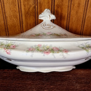 Puede incluir: Un plato de servir de porcelana blanca con tapa, con un diseño floral con rosas rosas y hojas verdes. El plato tiene un borde dorado y un borde festoneado.