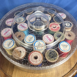 Może przedstawiać: Przezroczysty, okrągły zestaw do szycia z pokrywką, wypełniony zabytkowymi szpulkami nici w różnych kolorach. Szpulki są opisane tekstem i ułożone w okrąg. Zestaw stoi na drewnianej powierzchni.