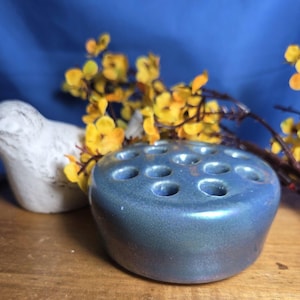 Puede incluir: Un ranero de flores de cerámica azul con múltiples agujeros, junto a una figura de pájaro blanco y flores amarillas. El ranero de flores tiene un diseño redondo y de perfil bajo con un acabado brillante.