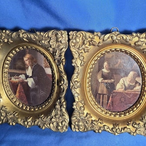 May include: Two antique oval framed prints with ornate gold-colored frames. The prints depict scenes of people in period clothing. The frames have a decorative, scrolled design. The background is a solid blue.