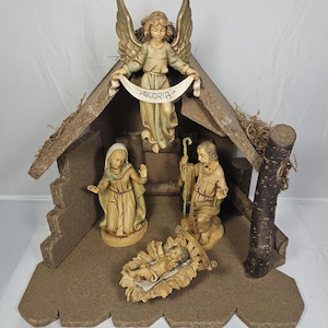 Puede incluir: Un pesebre de madera con un techo marrón, una base marrón y cuatro figuras. Las figuras son de María, José, el niño Jesús y un ángel que sostiene una pancarta que dice "Gloria".