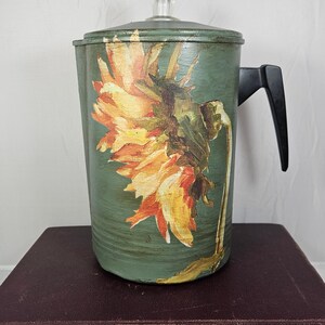 Könnte beinhalten: Eine grüne Kaffeekanne aus Metall mit einem Sonnenblumenmuster. Die Kanne hat einen schwarzen Griff und einen Glasdeckel.