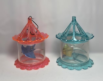 May include: Two decorative, clear plastic carousel-shaped containers. One is coral-colored, the other is teal. Each has a scalloped base and top, with a small pinwheel inside. A black string is attached to the coral container.