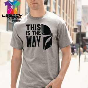 Puede incluir: Una camiseta gris con un gr&aacute;fico negro de un casco mandaloriano y el texto "This is the way".