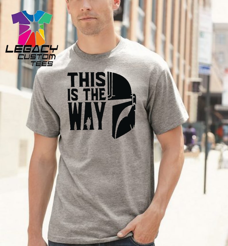 Puede incluir: Una camiseta gris con un gr&aacute;fico negro de un casco mandaloriano y el texto "This is the way".