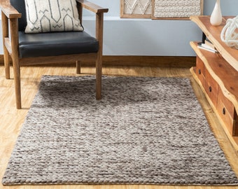 Puede incluir: Una alfombra de área texturizada marrón y beige con un aspecto tejido. La alfombra está en un ambiente de sala de estar con una silla de madera y un centro de entretenimiento de madera.