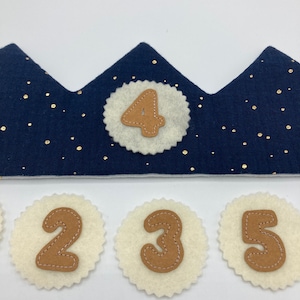 Könnte beinhalten: Eine blaue Filzkrone mit goldenen Glitzer und einem weißen Filzkreis mit der Zahl 4 in braunem Filz. Es gibt drei weitere weiße Filzkreise mit den Zahlen 2, 3 und 5 in braunem Filz.