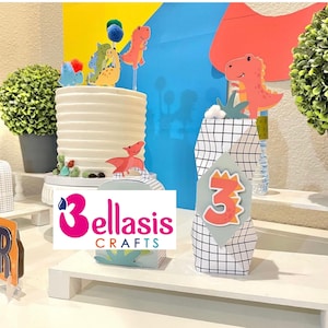 Puede incluir: Un pastel blanco con una superficie texturizada, decorado con coloridas decoraciones de dinosaurios. El pastel se encuentra sobre un soporte blanco, junto a una bolsa de regalo con un recorte del número 3. La escena se desarrolla sobre un fondo de formas azules y amarillas, con una pequeña planta en maceta.