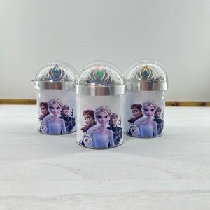 Puede incluir: Tres contenedores cilíndricos blancos con tapas plateadas y una cúpula de plástico transparente en la parte superior. Cada contenedor presenta una imagen colorida de personajes de la película Frozen, incluyendo Elsa, Anna, Kristoff y Olaf. Las tapas tienen un diseño de copo de nieve plateado.