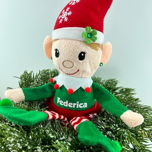 Può includere: Una bambola di elfo di peluche con un cappello rosso, un vestito verde e bianco e il nome "Federica" ricamato sul davanti. L'elfo è seduto su un letto di rami di pino verdi.