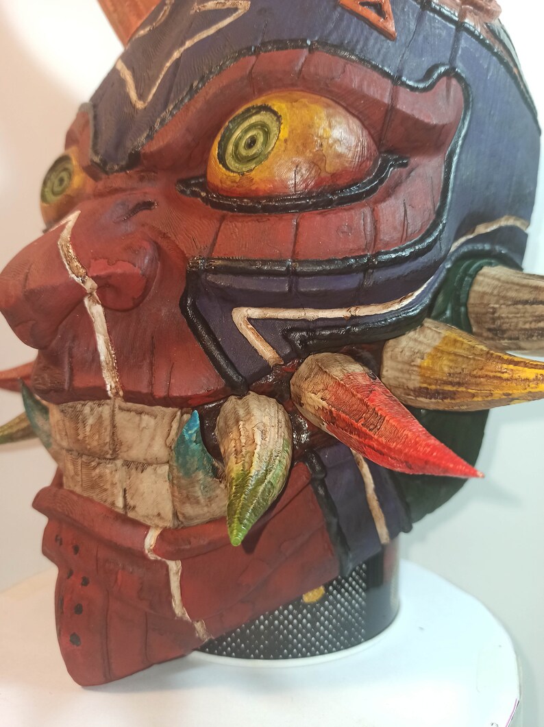 May include: A hand-painted, red, and brown mask with a detailed design. The mask has a large, yellow eye with a green spiral inside. The mask has a set of sharp, colorful teeth.