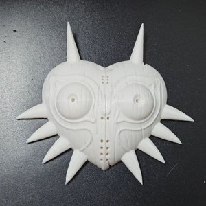 May include: A white plastic 3D printed mask with a heart shape and sharp points around the edges. The mask has two large eye holes and a detailed design.
