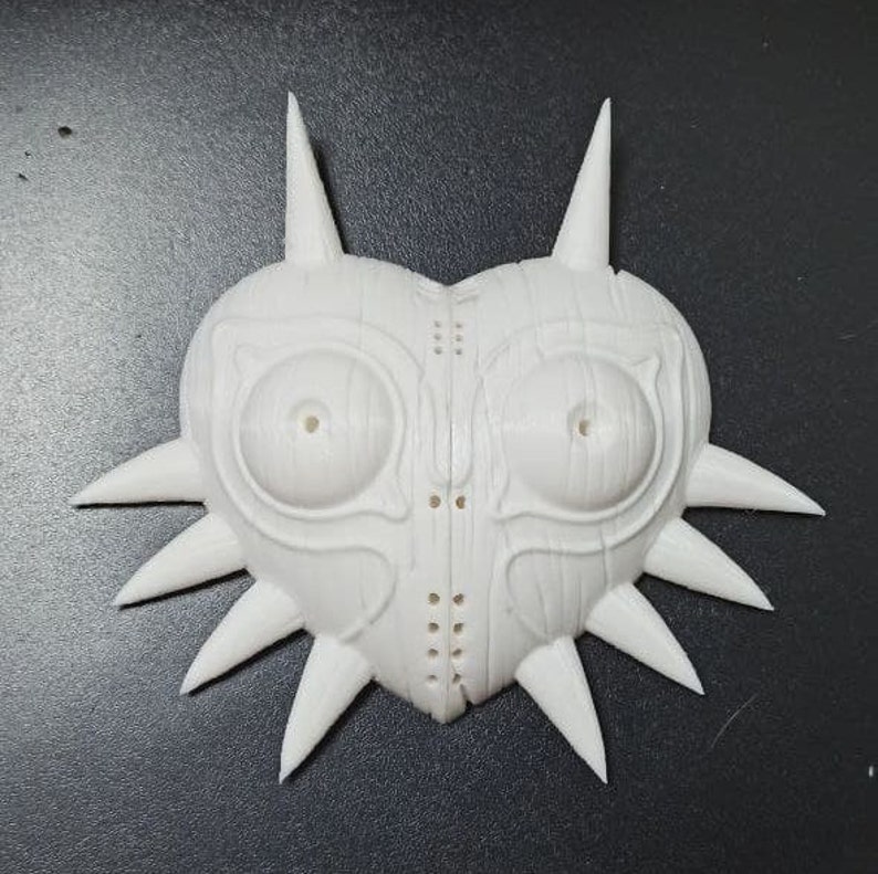 May include: A white plastic 3D printed mask with a heart shape and sharp points around the edges. The mask has two large eye holes and a detailed design.