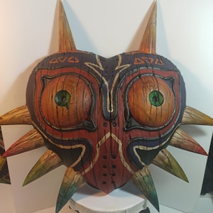 May include: A hand-painted wooden mask with a heart shape and sharp points. The mask is painted in shades of red, brown, green, and black. It has two large eyes with green pupils and a detailed design.