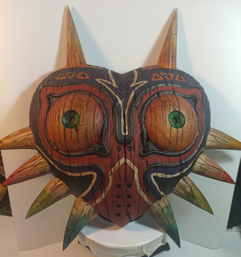 May include: A hand-painted wooden mask with a heart shape and sharp points. The mask is painted in shades of red, brown, green, and black. It has two large eyes with green pupils and a detailed design.
