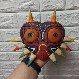 May include: A hand holds a large, heart-shaped mask with a purple, red, and orange design. The mask has two large, orange eyes with green pupils and a series of sharp, wooden spikes around the edges.
