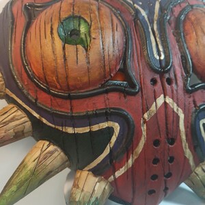 May include: A wooden mask with a red, black, and green paint design. The mask has a heart shape with sharp points around the edges. The mask has a large eye with a green iris and a black pupil.