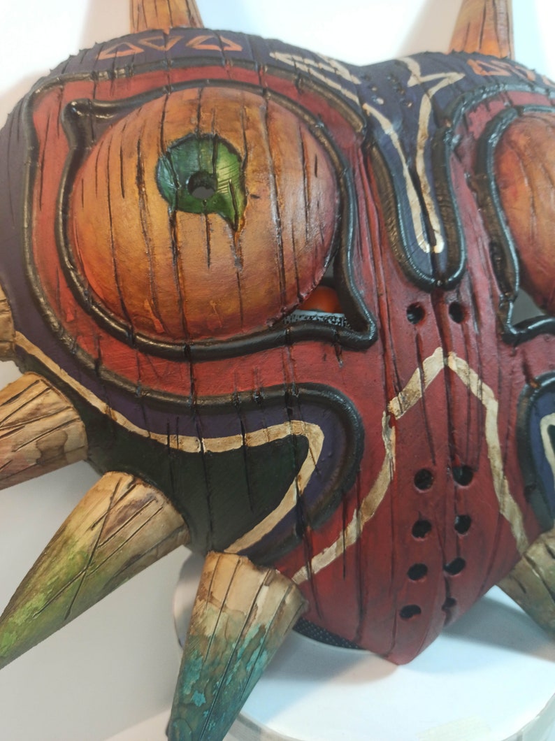 May include: A wooden mask with a red, black, and green paint design. The mask has a heart shape with sharp points around the edges. The mask has a large eye with a green iris and a black pupil.