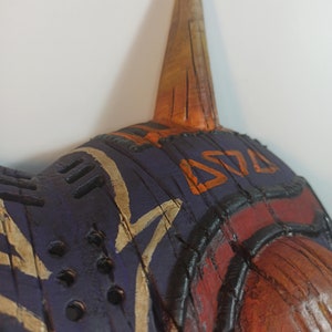 May include: A wooden mask with a purple, orange, and brown paint design. The mask has a long, pointed horn on top and a series of holes around the edges.
