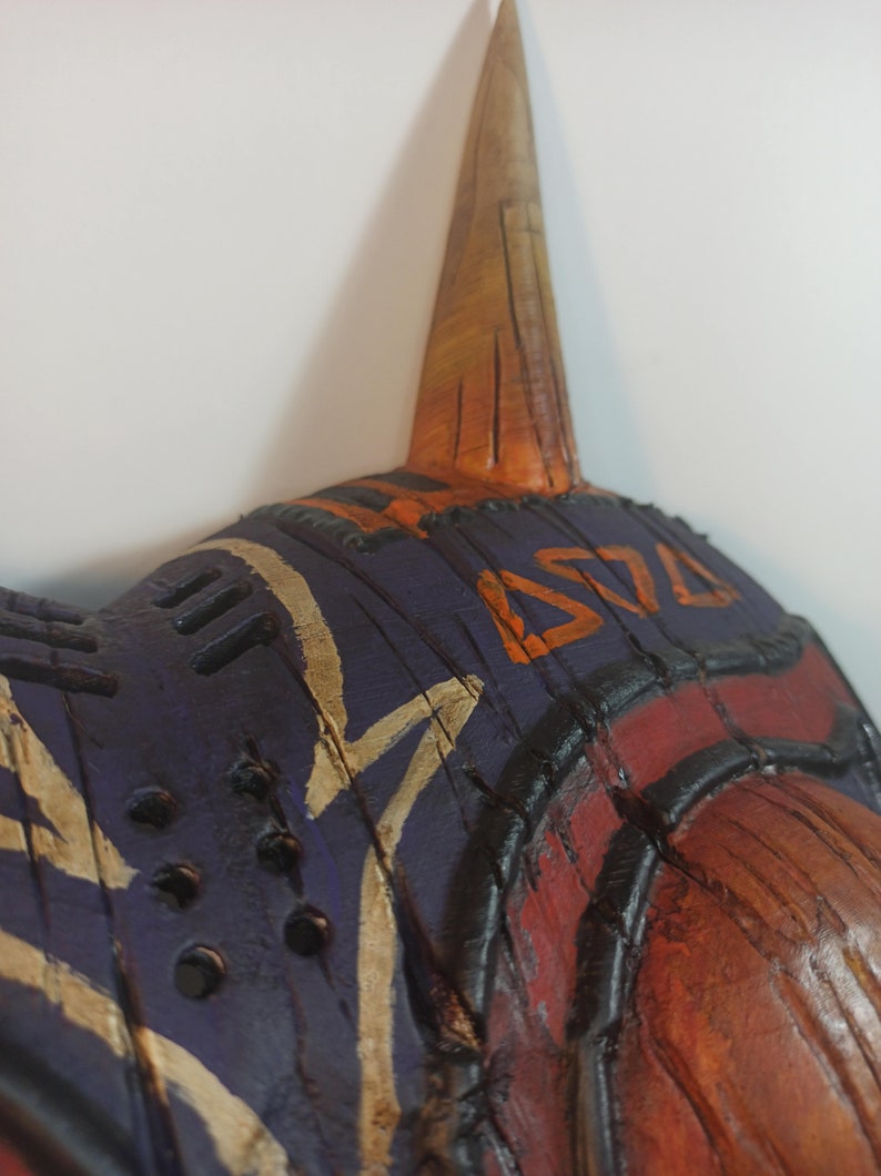 May include: A wooden mask with a purple, orange, and brown paint design. The mask has a long, pointed horn on top and a series of holes around the edges.