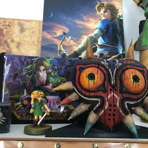 May include: A large, intricately carved wooden mask with red, orange, and blue paint. The mask has two large, round eyes and a sharp, pointed nose. The mask is on a black stand and is surrounded by other Zelda-themed items.