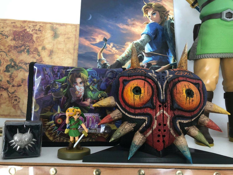 May include: A large, intricately carved wooden mask with red, orange, and blue paint. The mask has two large, round eyes and a sharp, pointed nose. The mask is on a black stand and is surrounded by other Zelda-themed items.