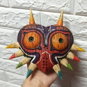May include: A hand-painted wooden mask with a heart shape and sharp points. The mask is painted in shades of red, orange, yellow, green, blue, and purple. It has two large eyes with green pupils and a detailed design.