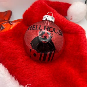May include: A red glitter Christmas ornament with the text "HELL HOUSE" and a black graphic. The ornament is resting on a red Santa hat with a white trim and pom-pom. The ornament has a silver metal cap.