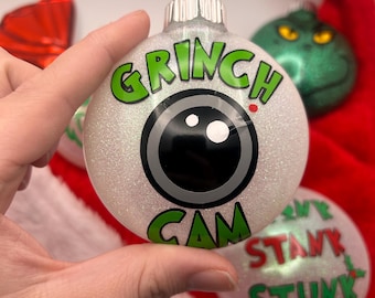 Puede incluir: Una bola de Navidad blanca brillante con las palabras "GRINCH CAM" en letras verdes. El adorno presenta un diseño de ojo negro y gris. Otros adornos y un gorro de Papá Noel están en el fondo.