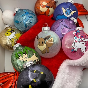 May include: A collection of glittery Christmas ornaments featuring various Pokémon characters. The ornaments are round, with silver metal caps, and are displayed on a red and white surface. The Pokémon designs include Eevee, Vaporeon, and others.