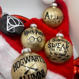 May include: A collection of Christmas ornaments with Harry Potter-themed designs. The ornaments are gold glitter, black, and silver. The text on the ornaments includes "Always", "Harry Potter", "Hogwarts Alumni", and "I Solemnly Swear That I am up to no good".