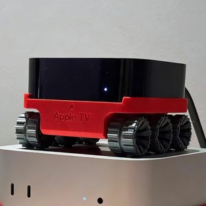 May include: A black Apple TV device sits atop a red base with six black wheels, resembling a small vehicle. The red base has the words "Apple TV" printed on it. The device is placed on a silver and white base.
