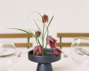 May include: A black, pedestal-style bowl holds a floral arrangement. The arrangement features green stems and leaves, with several maroon and white speckled flowers. Two clear, round glasses are on either side of the bowl.