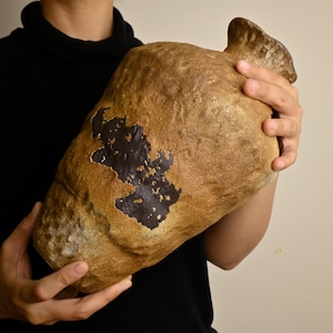 May include: A large, textured, tan-colored ceramic vessel with a wide, rounded body and a narrow neck. The surface features a prominent, irregular patch of dark brown, smooth glaze that contrasts with the rough, matte finish of the clay body.