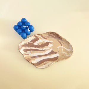 May include: A blue cube-shaped bubble candle with a white wick positioned next to an irregular, hand-formed ceramic tray featuring a marbled pattern of tan, brown, and white swirls across its smooth, glossy surface.