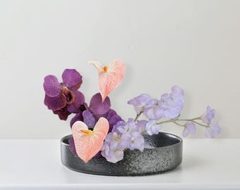 May include: A floral arrangement featuring purple orchids, peach anthuriums, and lavender sweet peas in a dark gray ceramic bowl. The flowers are arranged in a minimalist style, creating a visually appealing display.