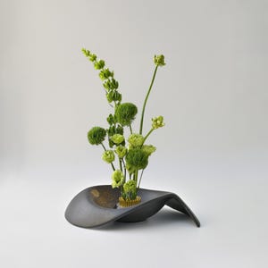 May include: A dark grey, asymmetrical ceramic bowl with a textured finish, holding a gold-toned metal kenzan flower frog. A collection of light green flowers and tall, slender green stems are arranged vertically within the floral frog.