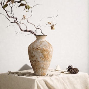 May include: A tall, textured beige vase with a dark rim holds branches with dried leaves. The vase sits on a linen-covered table, accompanied by a pine cone and scattered leaves. The neutral tones create a natural, earthy aesthetic.