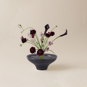 Puede incluir: Un jarrón de cerámica gris oscuro, de perfil bajo, con un arreglo floral. El arreglo presenta rosas de color burdeos intenso, calas y delicadas flores moradas y blancas. El jarrón tiene forma de cuenco ancho y poco profundo.