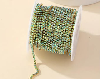 May include: A spool of iridescent green rhinestone chain. The chain is made of small, square rhinestones that are set in a gold metal setting. The chain is wound around a white spool.