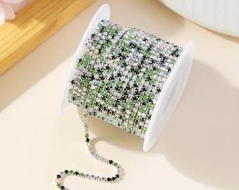 May include: A spool of clear, green, and silver rhinestone chain. The chain is rolled up on a white spool and a section of the chain is hanging down.