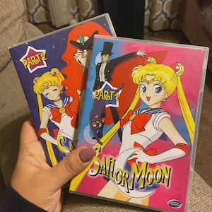 May include: Two VHS tapes of the anime Sailor Moon. The first tape is labeled "Part 1" and features a black-clad figure with a black cat. The second tape is labeled "Part 2" and features a blonde-haired girl with a blue and red outfit. Both tapes have a pink background with the title "Sailor Moon" in white.
