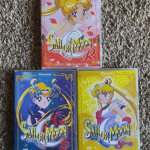May include: Three DVD cases for the Sailor Moon anime series. The cases are blue, yellow and red with the title "Sailor Moon" and the text "The Movie" on each. The cases are all in the same style with a picture of Sailor Moon on the front.
