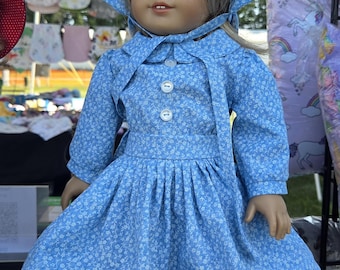 Könnte beinhalten: Eine Puppe mit blondem Haar und blauen Augen trägt ein hellblaues Kleid mit kleinem weißen Blumenmuster, langen Ärmeln, Bubikragen und drei weißen Knöpfen. Die Puppe trägt dazu eine passende, unter dem Kinn gebundene Haube mit breiter Krempe.