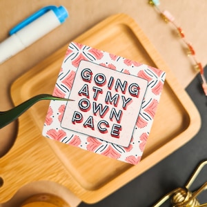 A glossy square shaped sticker with lettering saying "going at my own pace" in all caps