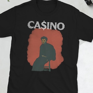 May include: Black t-shirt with the word "CASINO" in white rhinestone-style letters. The shirt features a graphic of a person in a dark coat sitting on a stool against a red background.