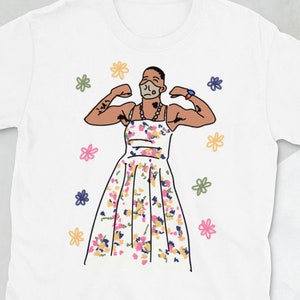 May include: White t-shirt featuring a graphic of a person wearing a floral dress and a face mask, flexing their biceps. The graphic is surrounded by small floral designs.