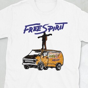 May include: A white short-sleeve t-shirt featuring a graphic illustration of an orange van with a person standing on its roof. Above the van, the text "FREE SPIRIT" is printed in a bold, dark blue, handwritten-style font.
