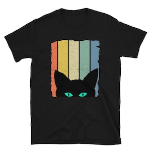 May include: Black t-shirt with a retro-style design of a cat peeking out from behind colorful stripes. The cat has green eyes.